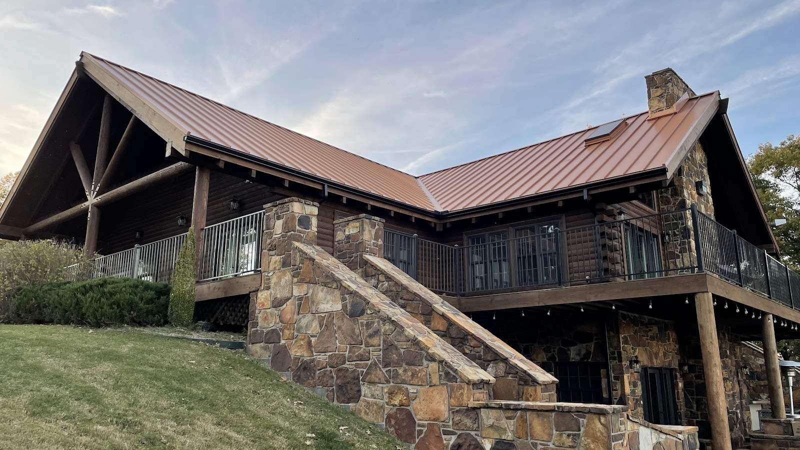A stained log-sided home with a standing-seam metal roof and a stone entry stair, photographed from the lawn below.