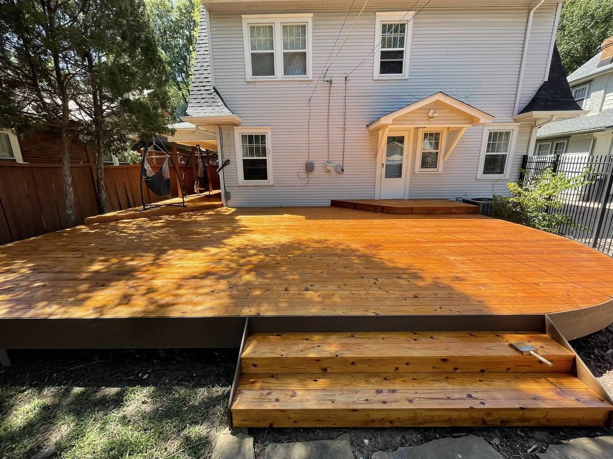 A wide cedar deck with two steps down to the lawn, stained a warm amber, behind a white two-story house.