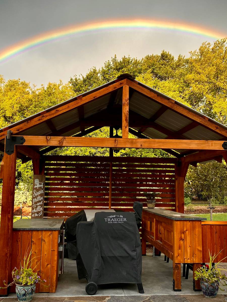 A timber gazebo with slatted walls and a grill station, stained a warm orange-brown, under a rainbow.