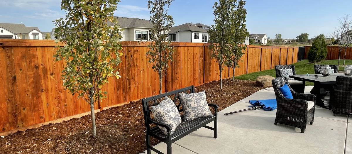 A reddish-brown stained wood privacy fence behind a concrete patio with outdoor seating and young trees.